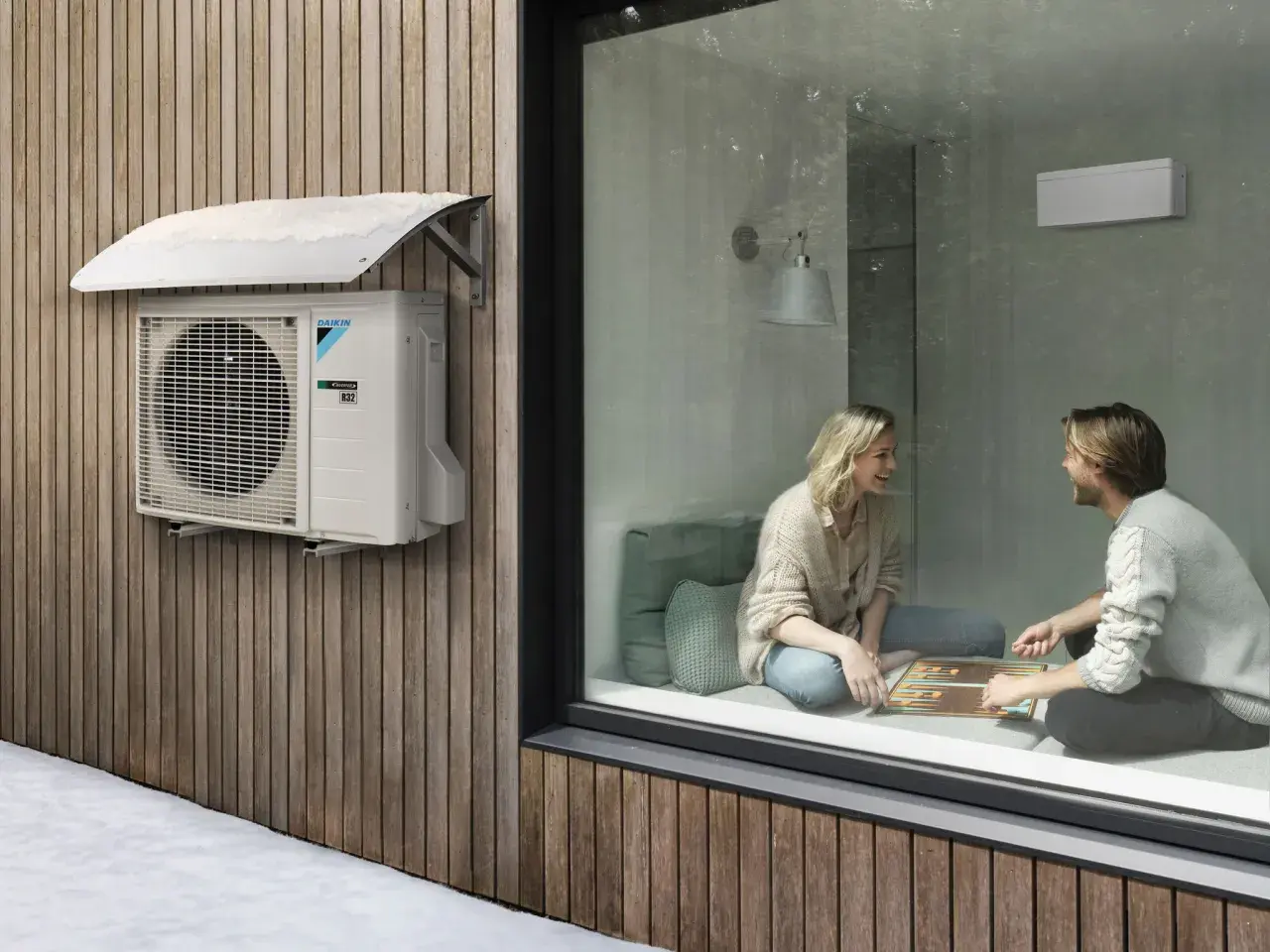 Unidad exterior de aire acondicionado Daikin instalada con la distancia adecuada a la pared de madera. Dentro, una pareja juega.