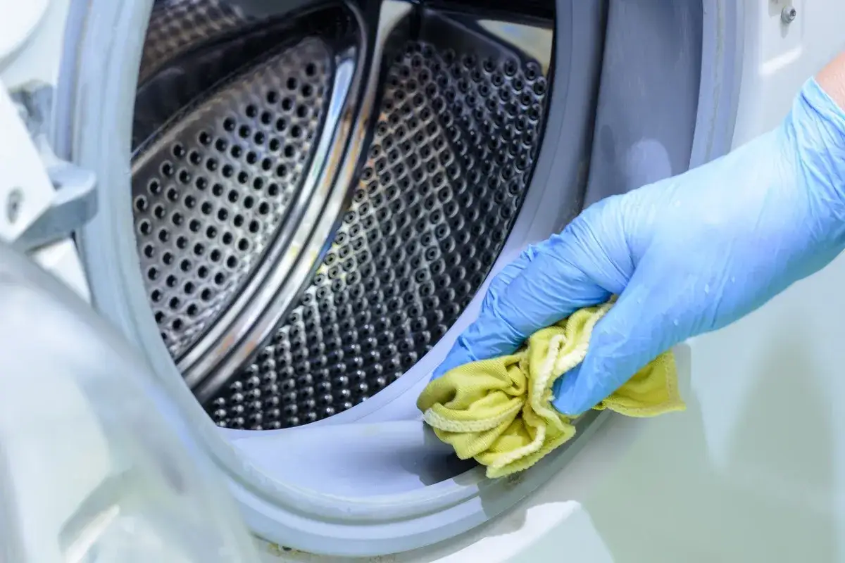 Mano con guante azul limpiando la goma de la lavadora con un pa&ntilde;o amarillo. Aprende como quitar el mal olor de la lavadora.