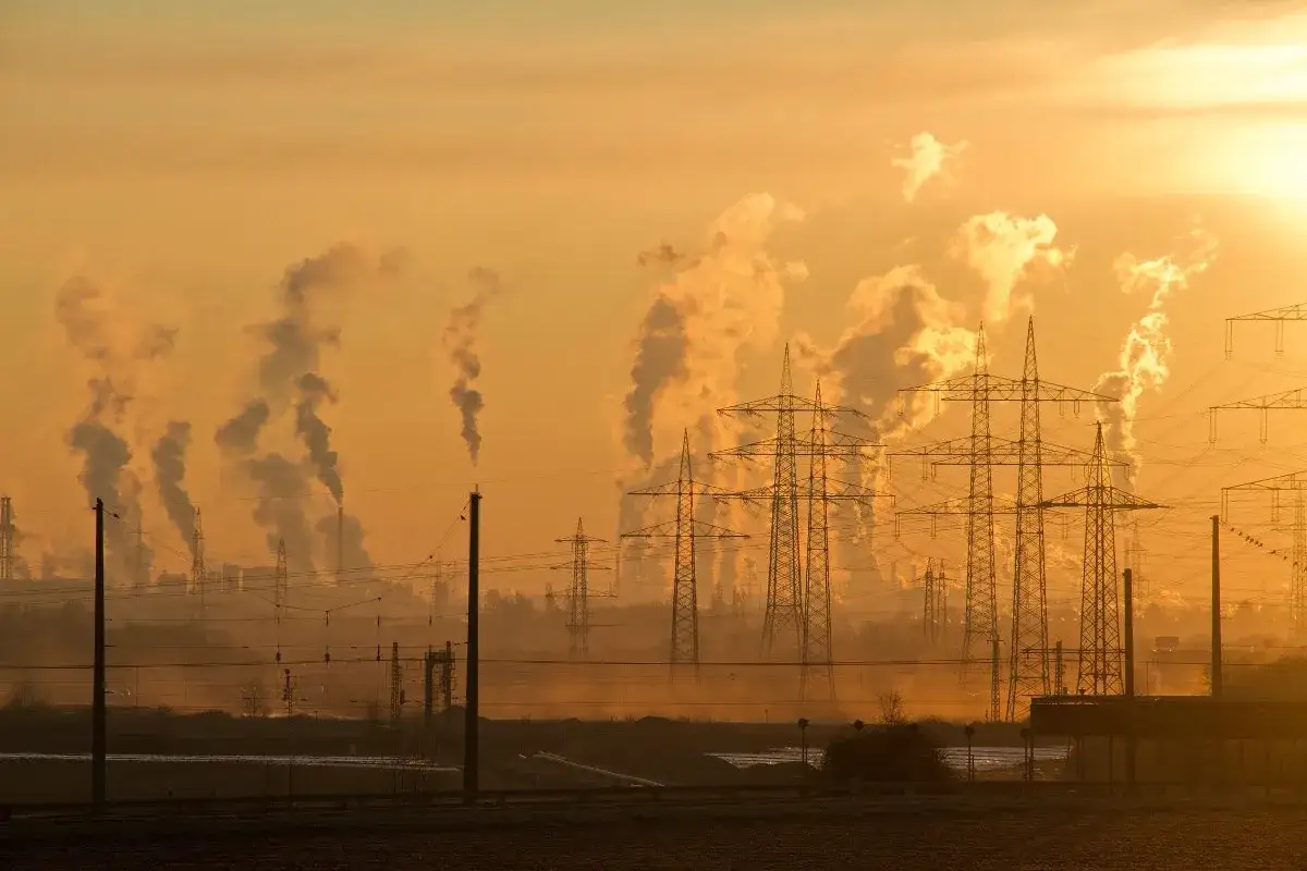 Siluetas de chimeneas industriales emitiendo humo, creando una atm&oacute;sfera de nox contaminante. Torres el&eacute;ctricas se alzan contra el cielo anaranjado del atardecer.