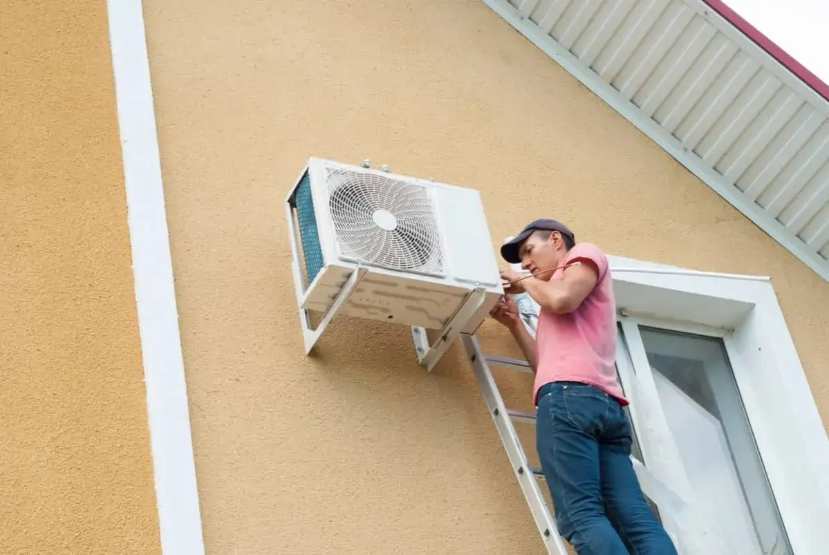 T&eacute;cnico en escalera instala unidad exterior de aire acondicionado inverter en pared de casa.