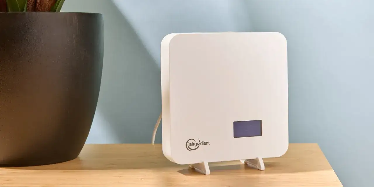 Dispositivo blanco mide la calidad del aire iaq. Junto a una maceta oscura, en una mesa de madera.