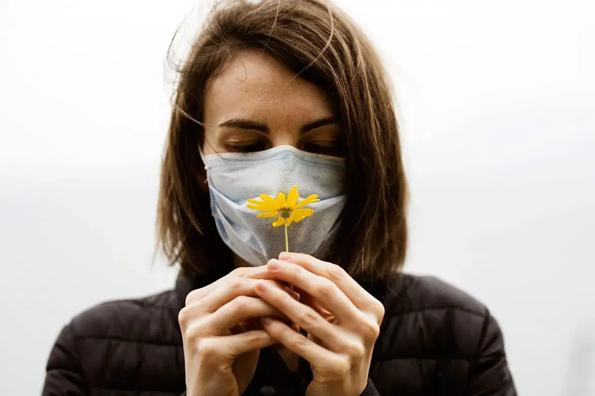 Mujer con mascarilla quirúrgica para que sirve, huele una flor amarilla.