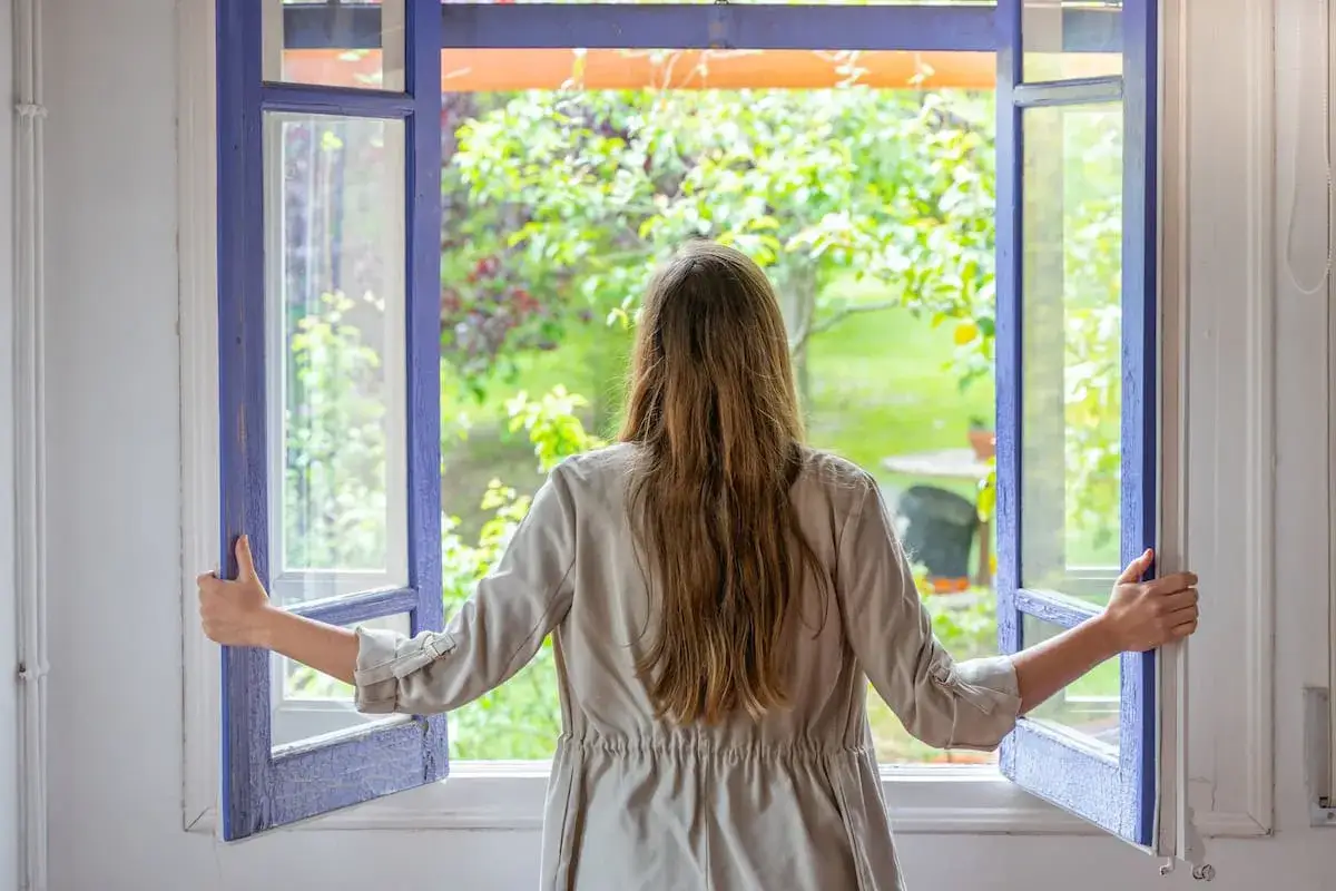 Mujer abriendo ventana para purificar el aire de casa de forma natural. El aire fresco del jard&iacute;n entra en la habitaci&oacute;n.
